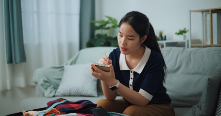 Selective focus of Asian teenager woman sitting on sofa making checklist of things to pack for travel, Preparation travel suitcase at home. - 772699488