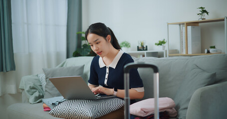 Portrait of Asian teenager woman sitting on sofa using laptop for prepare booking hotel and airplane ticket for travel. backpacker travel concept. - 772699616