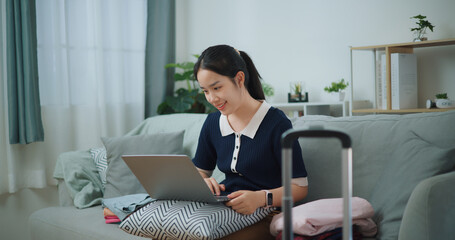 Portrait of Asian teenager woman sitting on sofa using laptop for prepare booking hotel and airplane ticket for travel. backpacker travel concept. - 772699631