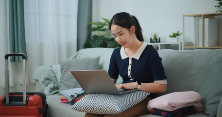 Portrait of Asian teenager woman sitting on sofa using laptop for prepare booking hotel and airplane ticket for travel. backpacker travel concept. - 772699673