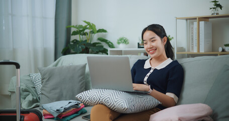Portrait of Asian teenager woman sitting on sofa using laptop for prepare booking hotel and airplane ticket for travel. backpacker travel concept. - 772699698