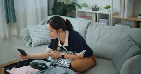 High angle view of Asian teenager woman sitting on sofa making checklist of things to pack for...