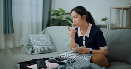 Portrait of Asian teenager woman sitting on sofa making checklist of things to pack for travel, Preparation travel suitcase at home.