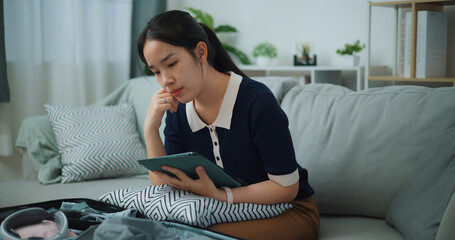 Side view of Asian teenager woman sitting on sofa planning vacation trip and searching information on digital tablet, travel and lifestyle.