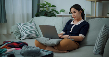 Portrait of Asian teenager woman sitting on sofa using laptop for prepare booking hotel and airplane ticket for travel. backpacker travel concept. - 772700273