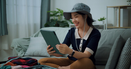 Portrait of Asian teenager woman sitting on sofa video call with friend while packing suitcase luggage for travelling, backpacker travel concept. - 772700433