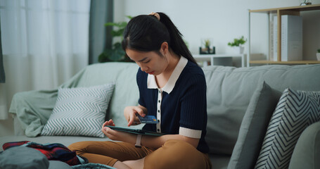 Portrait of Asian teenager woman sitting on sofa holding credit card making online payment on digital tablet, Preparation travel suitcase at home. - 772700445