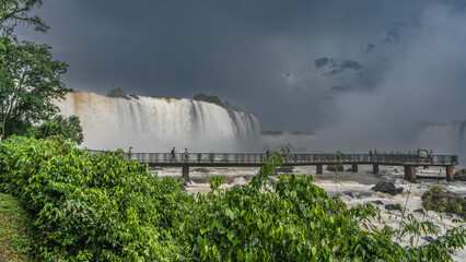 A pedestrian tourist bridge is laid over a stormy river. Tiny silhouettes of people are visible inspecting the waterfall. The spray rises into the sky like a cloud. Tropical vegetation. Iguazu Falls.