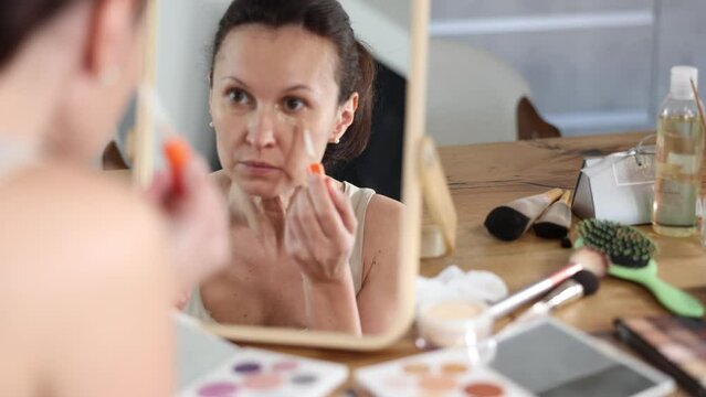 Serene confident adult woman applying under-eye concealer carefully in well-lit bedroom, sitting before wooden framed makeup mirror 