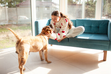 A beautiful young girl is wearing s white sweater with red hearts and jeans, sitting on the coach in living eoom against the windows  and hugging her animal friend.  Sharpey dog of a biege color