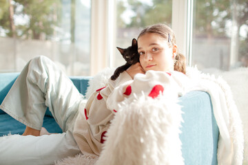 The girl holding the black cat. Friendship between the human and animal. The female dressed the white sweater with red heart. The portrait of young woman holding the kitten  rest in the blue sofa