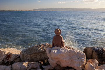 Back view. relaxed girl in a hat and coat sitting in front of the sea on the stones.