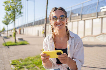 Positive hipster girl holds mobile phone, looks at cellphone screen, uses smartphone apps. girl with mobile phone on the street. 