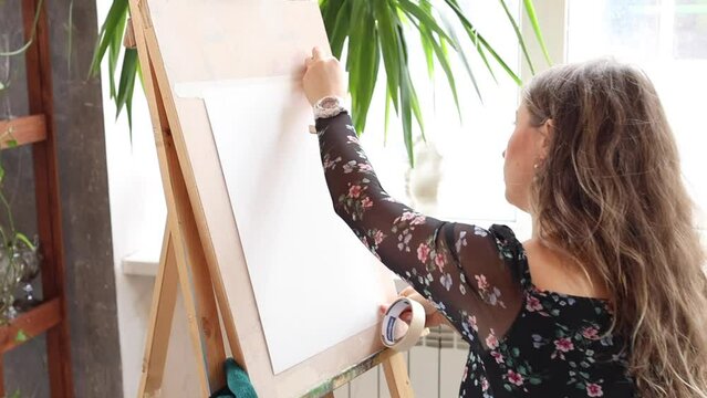 Beautiful young woman starting to paint in light art studio. Female artist sticking tape on paper and wooden easel. Blonde girl in black floral dress in creative process