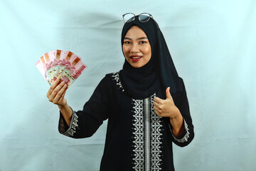 Smiling beautiful young Asian Muslim woman wearing hijab holding cash in rupiah banknotes in hand...