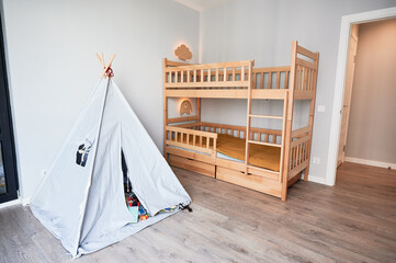 Fototapeta na wymiar Kid room with wooden bunk bed, wigwam and elegant minimalist interior design in apartment after renovation. Child bedroom with comfortable kid bed, laminate floor, white walls and open door.