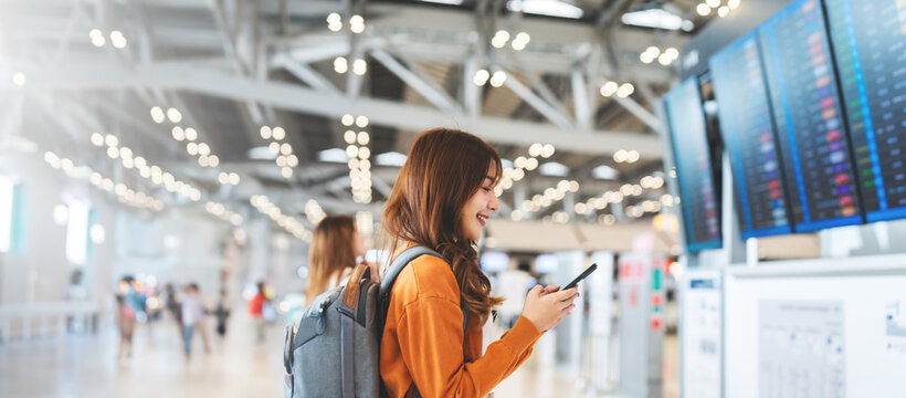 Young asian woman in international airport, using mobile smartphone and checking flight at the flight information board