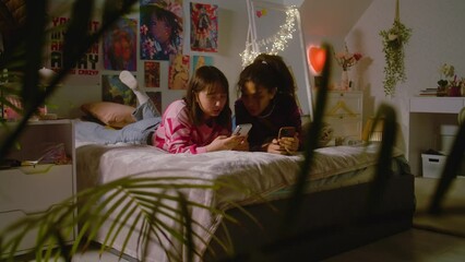 Two multiethnic best friends in the cozy bedroom laugh while scrolling social networks using mobile phone. Happy teen girls spend leisure time at home lying on the bed. Besties having fun together.