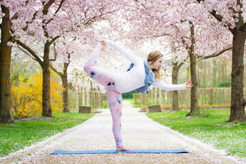 A young woman practices yoga against the backdrop of a spring cherry blossom alley. Tuladandasana, bow pose. Concept of mental, physical health and balance.