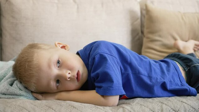 Blonde-haired blue-eyed child in casual clothes lies on the sofa at home. Portrait of a lying lazy bored preschooler boy. The kid is watching TV. Domestic life of real people.