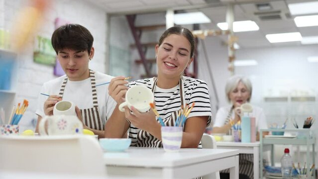 Young couple joyfully talking and working together on ceramic pottery, enjoying creative process while painting mugs in light workshop