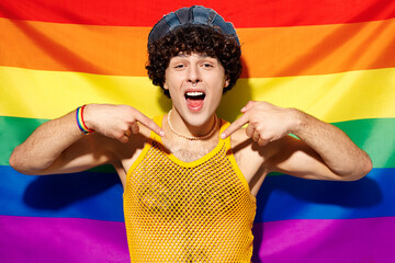 Young excited fun happy gay Latin man wears mesh tank top hat clothes point index fingers on himself stand on striped rainbow flag background studio portrait. Pride day June month love LGBT concept.