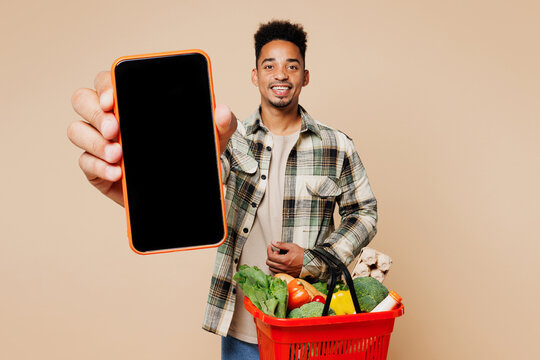 Young man wear grey shirt hold red basket bag with food products use close up blank screen workspace area mobile cell phone isolated on plain beige background Delivery service from shop or restaurant