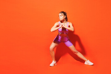 Full body young fitness trainer instructor woman sportsman wearing top shorts purple clothes train in home gym do squats look aside isolated on plain orange background. Workout sport fit abs concept.