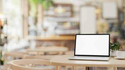 A white-screen laptop computer mockup on a wooden table in a contemporary cosy coffee shop. - obrazy, fototapety, plakaty
