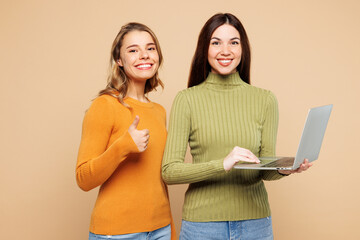 Young friends two IT women they wears orange green shirt casual clothes together hold use work on laptop pc computer show thumb up isolated on plain pastel light beige background. Lifestyle concept.