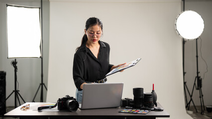 Female photographer is focusing on checking photoshoot details on a clipboard, working in her studio - obrazy, fototapety, plakaty