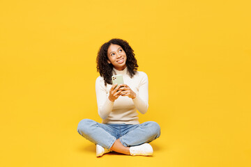 Full body fun little kid teen girl of African American ethnicity wear white casual clothes sits hold use mobile cell phone look aside isolated on plain yellow background. Childhood lifestyle concept.