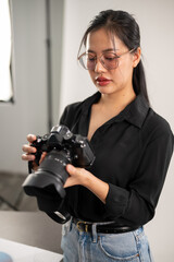 A confident, experienced Asian female photographer is adjusting her DSLR camera, preparing photoshoot equipment, and working in the studio.