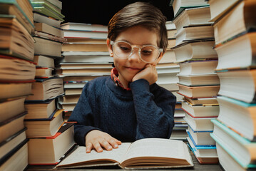 Cute little pupil boy in glasses reading book in library between stacks of books