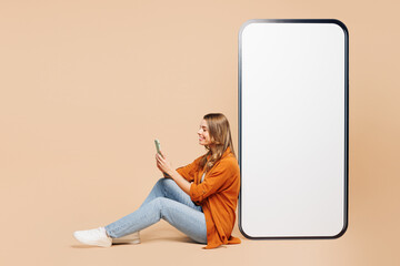 Full body side profile view young woman she wear orange shirt casual clothes sit near big huge blank screen mobile cell phone with mockup area use smartphone isolated on plain pastel beige background - 772798212