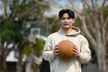 Handsome man holding ball with both hands and smiling at camera, standing on the playground