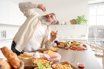 Overweight mature man at table full of unhealthy food in kitchen. Overeating concept