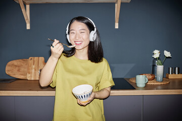 Flash portrait of joyful young Asian girl wearing yellow-green T-shirt and headphones standing against kitchen counter smiling at camera while eating