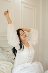 A beautiful Asian woman in long dress pajamas is stretching her arms after waking up.