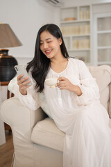 A beautiful Asian woman in long dress pajamas is using her smartphone and sipping tea on a sofa.