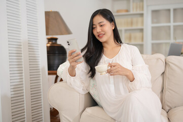 A beautiful Asian woman in long dress pajamas is using her smartphone and sipping tea on a sofa.