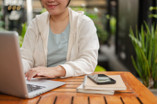 A cropped image of a happy Asian woman in casual wear is working on her laptop at an outdoor space.