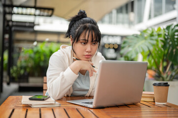 A serious, focused Asian woman is working remotely at an outdoor space, reading email on her laptop.