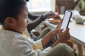 Fototapeta premium Side view portrait of teen African American boy using smartphone with blank white screen while sitting at kitchen table during breakfast