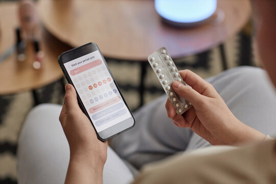 High angle view of unrecognizable young woman holding blister with birth control pills and checking period calendar on smartphone - Powered by Adobe