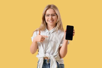 Fotobehang Cute little girl in eyeglasses pointing at mobile phone on yellow background © Pixel-Shot