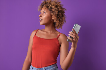 Young positive attractive African American woman teen with mobile phone in hand looks to side and opens mouth in surprise after seeing amazing offer for smartphone users stands in purple studio.