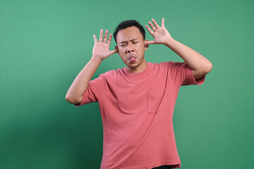 Portrait of silly Asian male making funny faces with hands on face and showing tongue