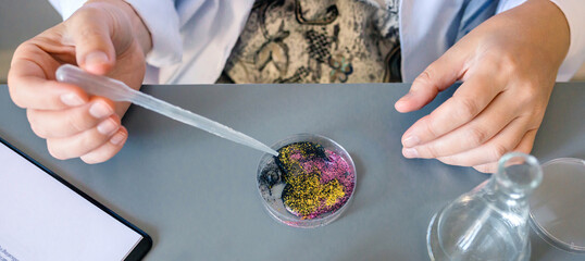 Banner of unrecognizable female scientist adding drop of liquid from from pipette over petri dish with mix of colorful glitters in environment research lab. Study of microplastics composition concept.