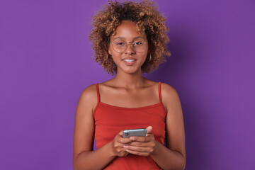 Young smiling beautiful African American woman teenager in glasses holds smartphone in hands and looks at camera offering you to download new application for online shopping stands in purple studio.